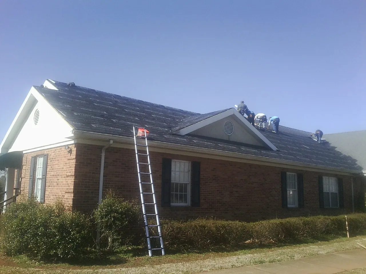 Crew tearing shingles off a brick ranch roof with a ladder set — Emergency Roof Repair work by our crew in Okauchee
