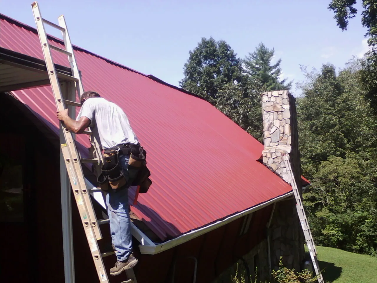 Roofer on a ladder beside a red metal roof and stone chimney — Soffit & Fascia Repair by Roofing Brothers in Okauchee